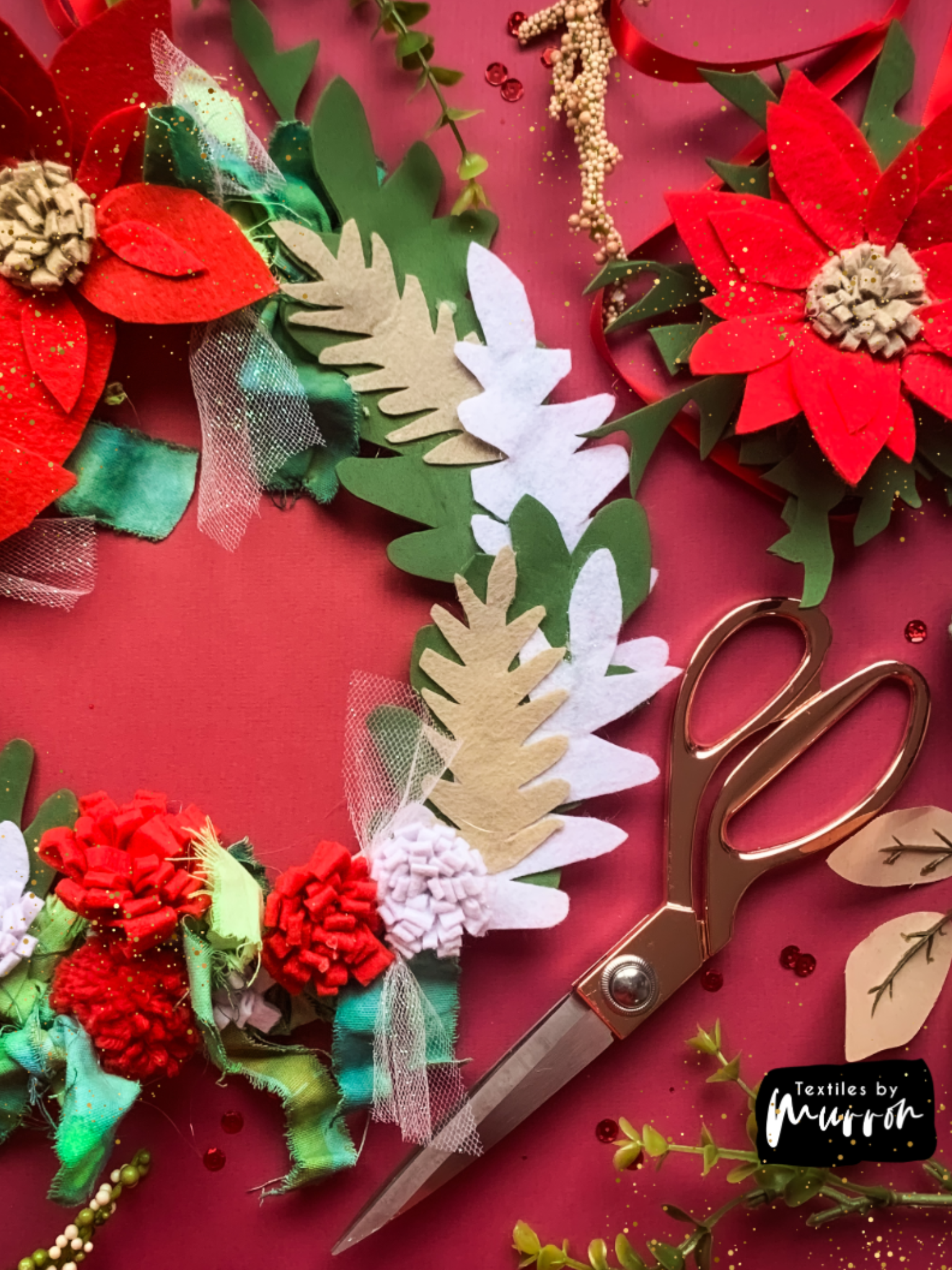 Red white and green textile wreath made in the festive workshop on a red table with gold scissors